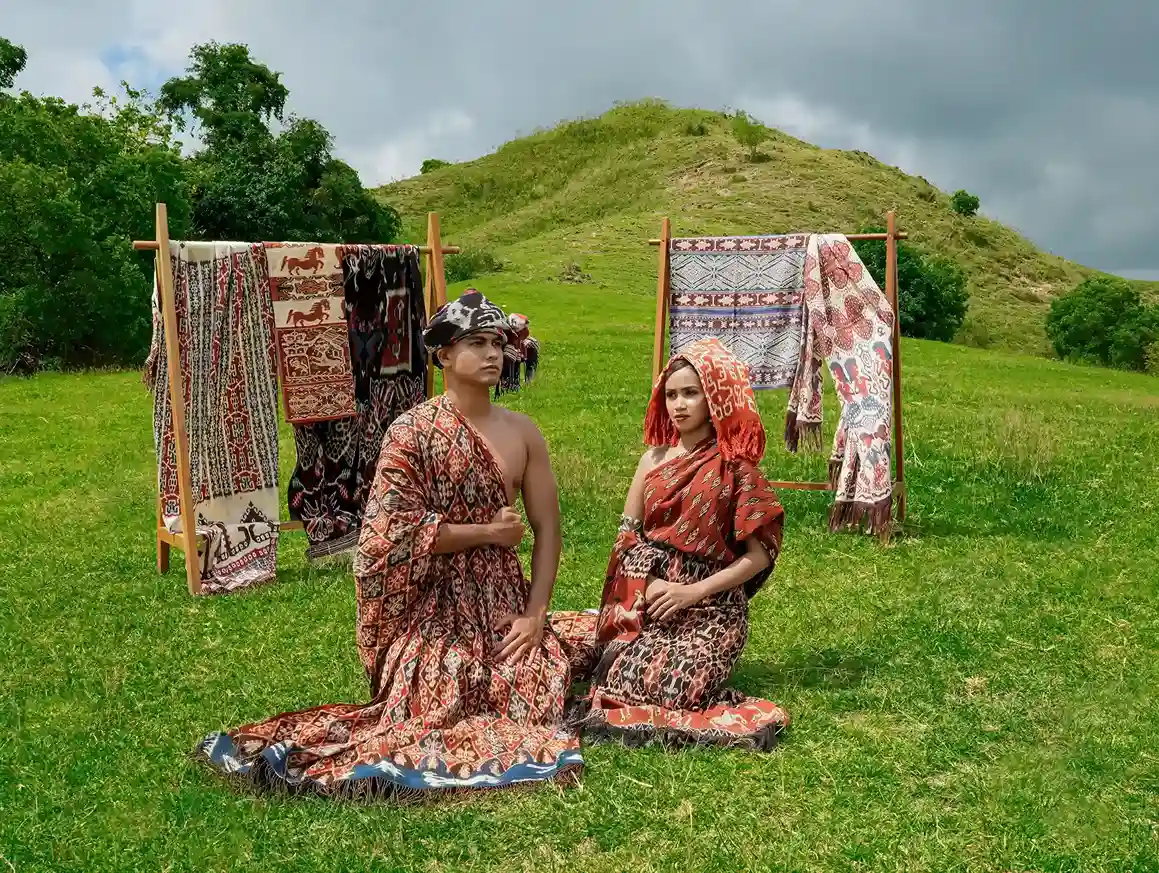 Handwoven Sumba ikat cloth displayed on wooden racks, with two people wearing traditional woven dress